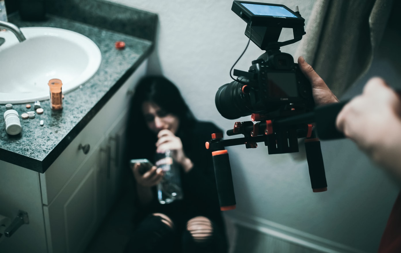 Mulher deitada em chão de banheiro, chorando, segurando uma garrafa de bebida alcóolica e digitando em seu celular. Em primeiro plano na imagem, alguém com uma câmera profissional filma a mulher.