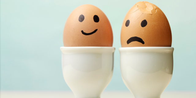 Dois ovos em taças brancas de porcelana. Um deles está com um rosto feliz desenhado na superfície, enquanto o outro está triste.