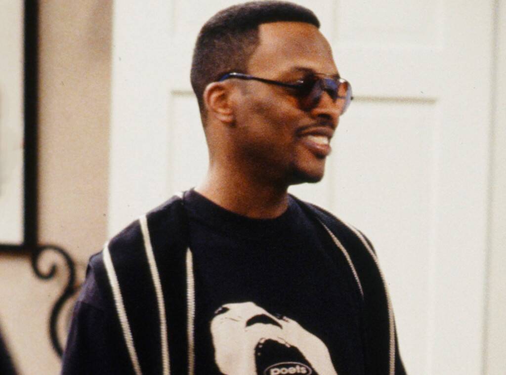Homem negro usando óculos de sol, blusa preta e com expressão sorridente.