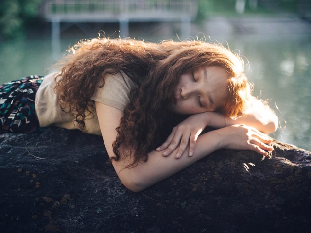 Mulher branca de cabelos longos e ruivos deitada com os olhos fechados.