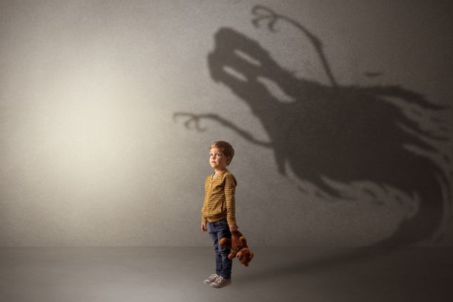 Garotinho em um ambiente vazio, segurando um ursinho de pelúcia. Na parede atrás dele, uma sombra assustadora simulando um fantasma.