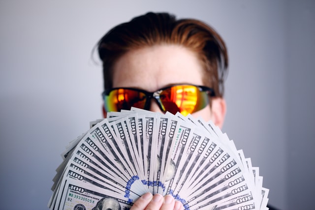 Homem branco de óculos espelhados, segurando leque de dólares em frente a boca.