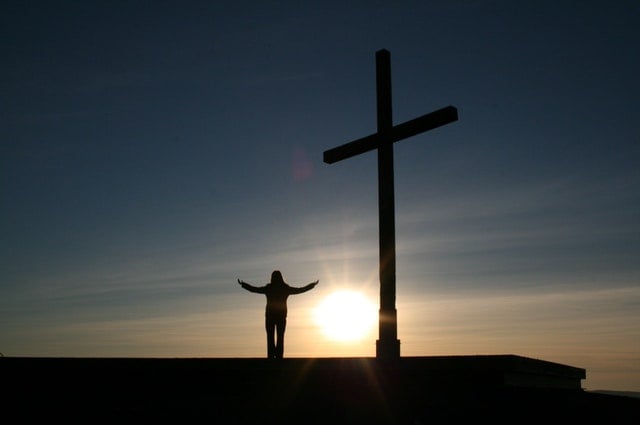 Silhueta de uma pessoa em pé, de braços abertos, ao lado de uma cruz
