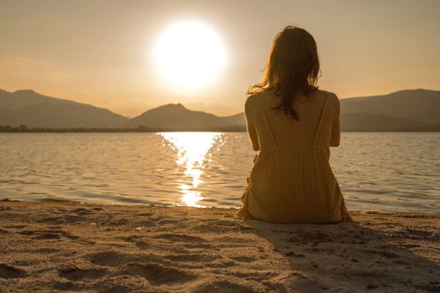 Mulher sentada na areia, sozinha, assistindo ao pôr-do-Sol.
