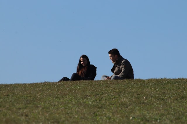 Homem e mulher brancos sentados na grama conversando.