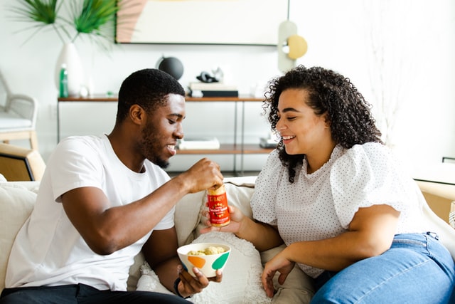 Homem e mulher negros sentados num sofá, comendo snacks.