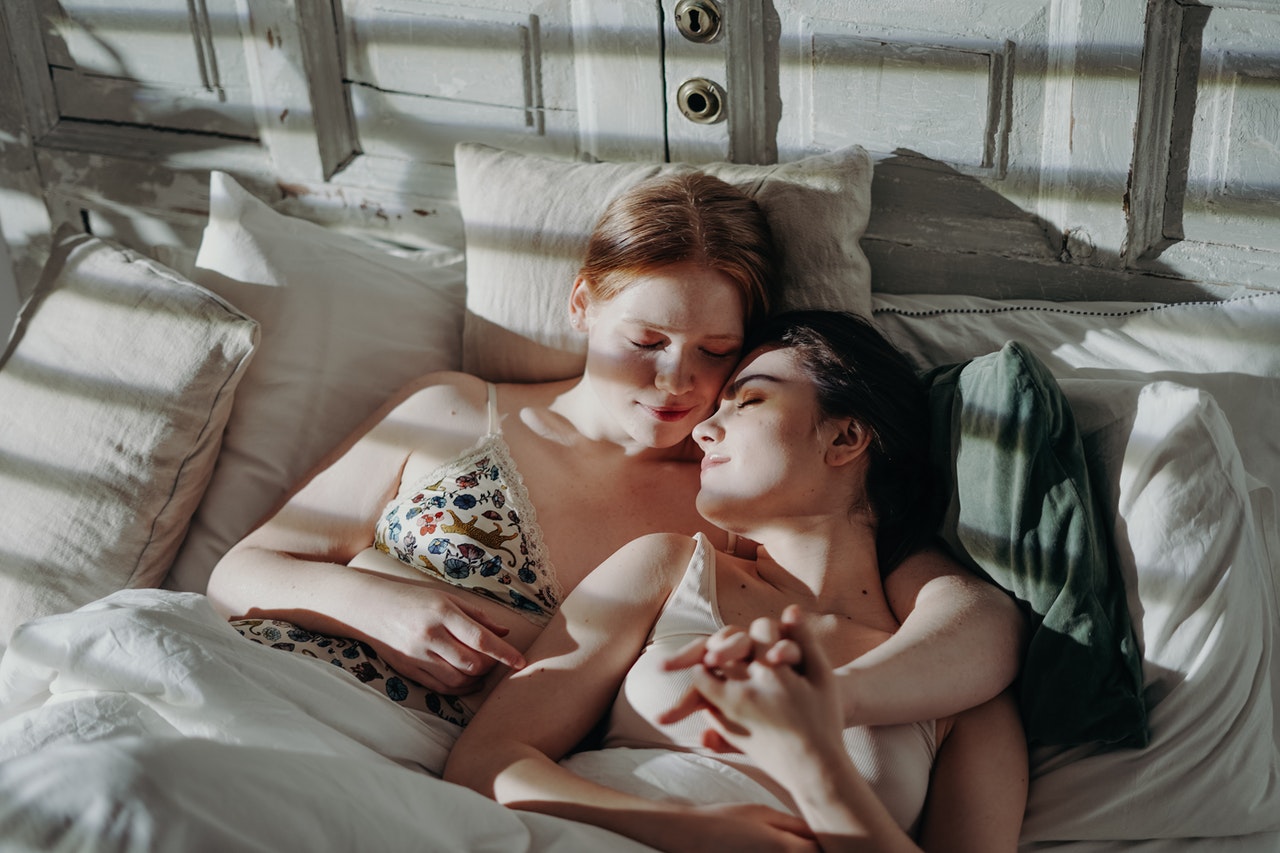 Duas mulheres deitadas em uma cama. Uma tem seu braço ao redor da outra, para que suas mãos se entrelacem.